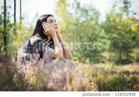 A young woman sits peacefully in a sunlit forest clearing, enjoying nature  119352930