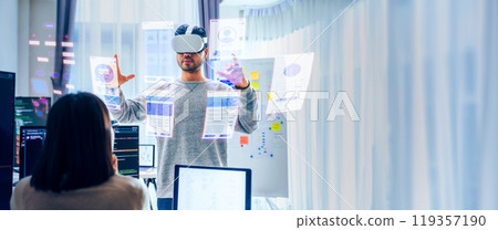Asian software developer wearing a virtual reality headset works on a VR project, with a colleague focused on her computer 119357190