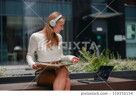 A Focused Young Woman Engrossed in Working Outdoors with Her Laptop and Headphones On A Focused Young Woman Engrossed in Working Outdoors with Her Laptop and Headphones On 119358154