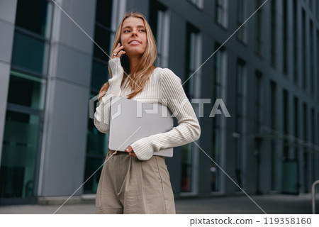 A young professional woman talks on her phone outdoors, enjoying a casual chic city vibe 119358160