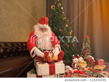 Santa Claus with gift box sitting on sofa at home near Christmas tree, presents, snowmen decorations and festive ornaments at night Santa Claus with gift box sitting on sofa at home near Christmas tree, presents, snowmen decorations and festive ornaments at night 119358395