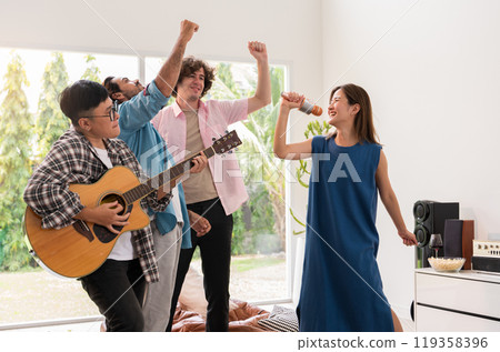 Friends enjoying lively music party at home. One playing guitar, one singing into microphone and other two dancing in living room. Friends enjoying lively music party at home. One playing guitar, one singing into microphone and other two dancing in living room. 119358396