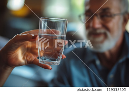 A close up of a hand holding a glass of water, with an elderly man smiling in the background A close up of a hand holding a glass of water, with an elderly man smiling in the background 119358805