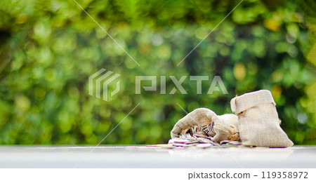 A burlap sack standing and another lying with coins spilling out onto a white surface, set against a blurred background of bushes, representing financial planning and budgeting. 119358972