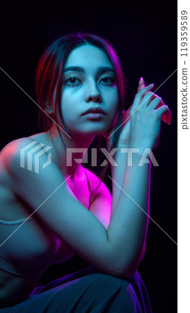 Portrait of beautiful young Korean girl posing against black background with blue and pink neon lights 119359589
