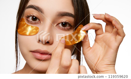 Korean woman applying golden under-eye patches, highlighting a skincare routine for reducing puffiness and brightening the under-eye area Korean woman applying golden under-eye patches, highlighting a skincare routine for reducing puffiness and brightening the under-eye area 119359620