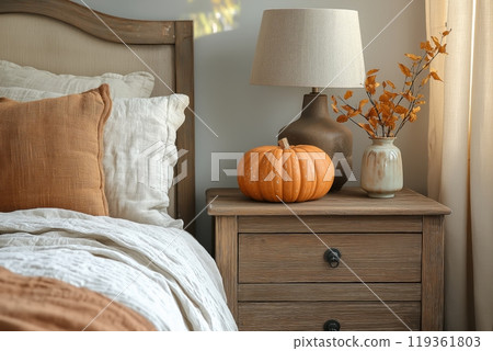 A wooden nightstand with a pumpkin, lamp, and vase of autumn leaves beside a cozy bed 119361803