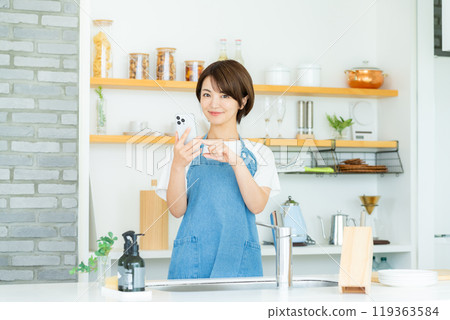 Smiling woman with smartphone in kitchen Smiling woman with smartphone in kitchen 119363584