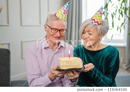 Make a wish. Family senior couple in party hat cap celebrating birthday anniversary together at home. Old woman blowing out burning candles on birthday cake. Old man wishes wife happy birthday Make a wish. Family senior couple in party hat cap celebrating birthday anniversary together at home. Old woman blowing out burning candles on birthday cake. Old man wishes wife happy birthday 119364500