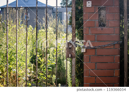Rusty old lock hangs on a chain at the gate on a red brick post on abandoned territory with greenery Rusty old lock hangs on a chain at the gate on a red brick post on abandoned territory with greenery 119366034