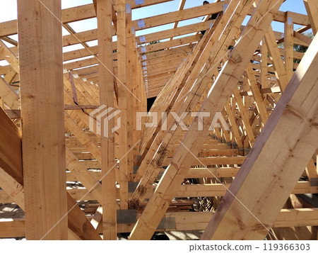 View through wooden trusses under construction, showcasing the symmetrical alignment and complex framework against the sky View through wooden trusses under construction, showcasing the symmetrical alignment and complex framework against the sky 119366303