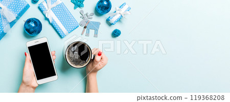 Top view of a woman holding a phone in one hand and a cup of coffee in another hand on blue background. Christmas decorations and toys. New Year holiday concept. Mockup 119368208