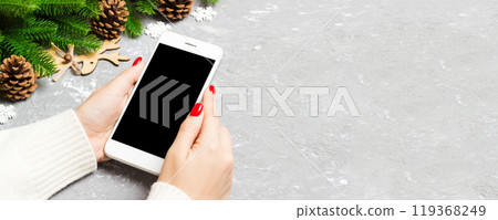 Top view of a woman holding a phone in her hand on cement New Year background made of fir tree and festive decorations. Christmas holiday concept. Mockup 119368249