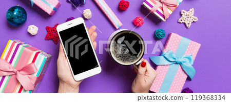 Top view of a woman holding a phone in one hand and a cup of coffee in another hand on purple background. Christmas decorations and toys. New Year holiday concept. Mockup Top view of a woman holding a phone in one hand and a cup of coffee in another hand on purple background. Christmas decorations and toys. New Year holiday concept. Mockup 119368334