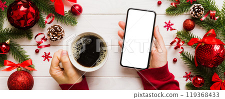 Above view of female hand holding smart phone with hot cup of coffee. Christmas decoration on colored background 119368433