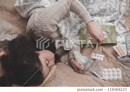 a sick girl in a sweater lying on the bed checks the temperature with a thermometer on the background of pills scattered on the blanket 119369223