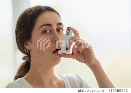 Asthma. Portrait of a young woman holding an inhaler to her mouth and looking at the camera. Blurry background. Copy space 119369232