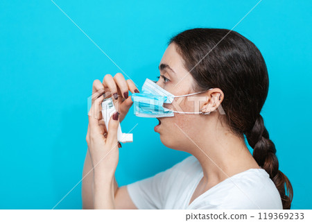 Asthma. Portrait of a young woman in a medical mask holding an inhaler to her mouth. Side view Asthma. Portrait of a young woman in a medical mask holding an inhaler to her mouth. Side view 119369233