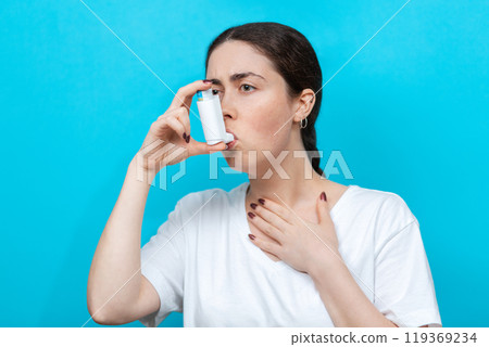 Asthma. Portrait of a young woman using an inhaler. Blue background Asthma. Portrait of a young woman using an inhaler. Blue background 119369234