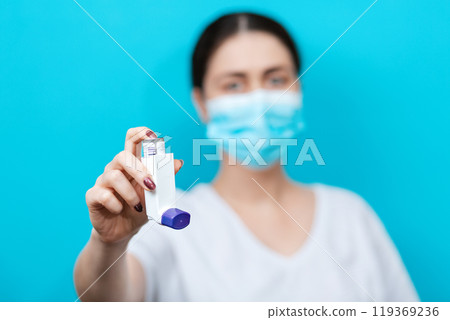 Blurry portrait of a female doctor in a medical mask showing an inhaler. Blue background. Copy space Blurry portrait of a female doctor in a medical mask showing an inhaler. Blue background. Copy space 119369236