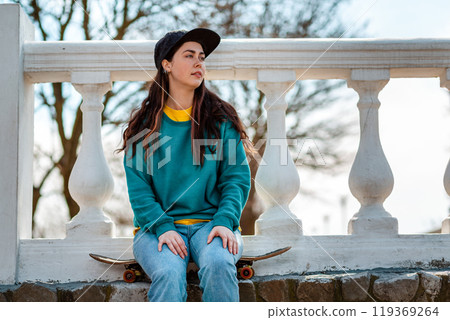 Portrait of a beautiful hipster teen girl sitting on a skateboard and looking into the distance. In the background is a white balustrade and tree.Concept of sports lifestyle and street culture Portrait of a beautiful hipster teen girl sitting on a skateboard and looking into the distance. In the background is a white balustrade and tree.Concept of sports lifestyle and street culture 119369264