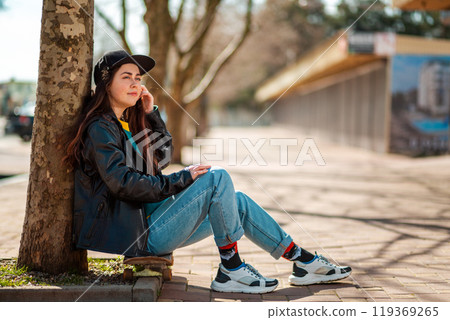 Portrait of a beautiful hipster teen girl sitting on a skateboard and looking into the distance. There's an alley with trees in the back. Copy space. Concept of sports lifestyle and street culture 119369265
