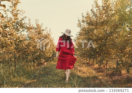 Portrait of a woman in the garden. A young and beautiful woman in a red dress and a straw hat walks through the orchard. Hot weather Portrait of a woman in the garden. A young and beautiful woman in a red dress and a straw hat walks through the orchard. Hot weather 119369271
