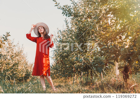 Portrait of a woman in the garden. A young and beautiful woman in a red dress and hat coquettishly takes off her hat. Copy space 119369272