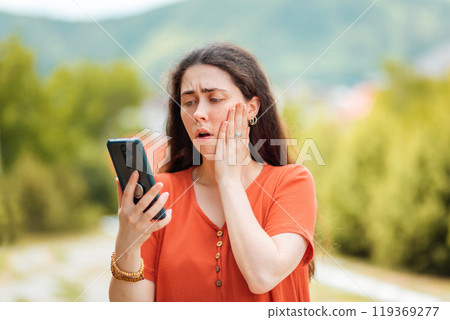 Portrait of a woman looking at the phone, clutching her face in fear. The Park is in the background. Concept of online fraud and fake news 119369277