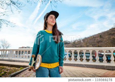 Portrait of a Young Caucasian woman holding a skateboard. In the background, the blue sky and the Boulevard. Copy space. Concept of sports lifestyle and street culture Portrait of a Young Caucasian woman holding a skateboard. In the background, the blue sky and the Boulevard. Copy space. Concept of sports lifestyle and street culture 119369278