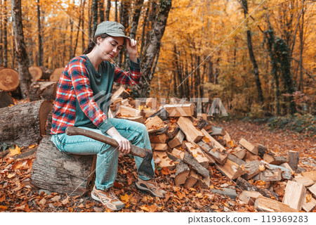 Preparation for the heating season. A young woman with an axe in her hands sits on a log and adjusts her cap on her head. In the background autumn forest 119369283