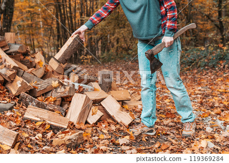 Preparation for the heating season. A young woman with an axe in her hands throws a log into a pile of logs. In the background autumn forest. Close up 119369284