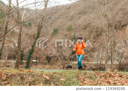Search With A Metal Detector. A young woman in an orange jacket is looking for lost artifacts in the Park area. Hobbies and activities 119369288