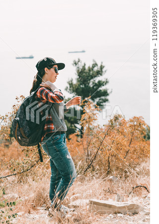 Tourism and outdoor activities. A woman in a cap and glasses, wearing a backpack. In the background the sea and trees. Vertical orientation Tourism and outdoor activities. A woman in a cap and glasses, wearing a backpack. In the background the sea and trees. Vertical orientation 119369305