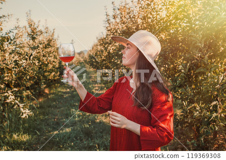 Wine tasting and relaxing in the garden. Young beautiful brunette in a red dress and hat holding a glass of wine. There are fruit trees in the background. Tint 119369308