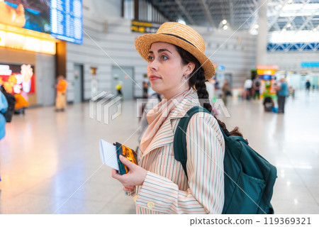Young Caucasian pretty woman in straw hat, wearing backpack, holds her passport to check in for a flight. Waiting at the train station 119369321