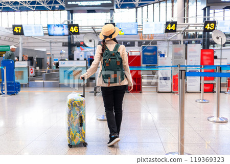 Elegant woman in a straw hat and a backpack rolls a suitcase through the airport. Rear view. Traveling by plane, check-in. Vacation concept Elegant woman in a straw hat and a backpack rolls a suitcase through the airport. Rear view. Traveling by plane, check-in. Vacation concept 119369323