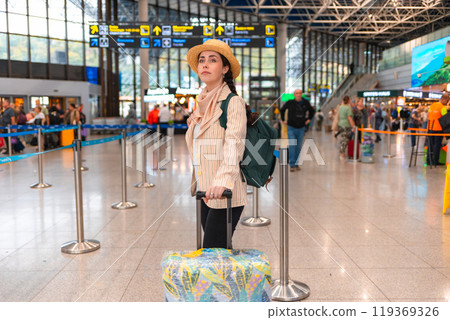 Elegant Caucasian woman wearing straw hat and backpack, holds suitcase and waiting in airport. Traveling by plane, check-in for a flight Elegant Caucasian woman wearing straw hat and backpack, holds suitcase and waiting in airport. Traveling by plane, check-in for a flight 119369326