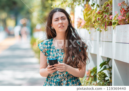 A woman with an indignant face holds a smartphone in her hands. In the background, Park street. Concept of communication and social networks 119369440