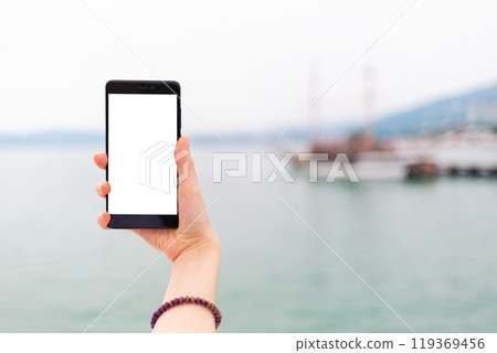 Mock up. The female hand holds the phone vertically. In the background, the sea and ships. Isolate 119369456