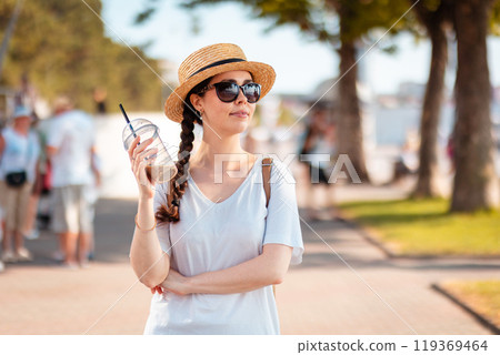 Portrait of young beautiful elegant Caucasian woman wearing straw hat strolls along the city boulevard holding drink in her hand. Blurred background. Concept of summer season vacation 119369464