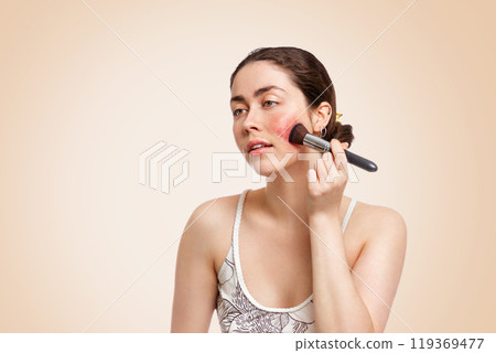 A young pretty woman applies makeup with a brush to the reddish area of inflammation. Beige background. Copy space. The concept of corrective makeup and rosacea A young pretty woman applies makeup with a brush to the reddish area of inflammation. Beige background. Copy space. The concept of corrective makeup and rosacea 119369477