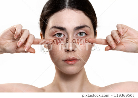 Close-up portrait of a young woman pointing at dark circles under her eyes. White background. Concept of skin care and combating the first signs of aging 119369505