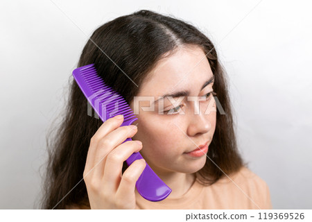 The woman is a brunette, parting her hair with a comb and showing the results of treatment for dandruff. White background. The concept of dandruff and pediculosis The woman is a brunette, parting her hair with a comb and showing the results of treatment for dandruff. White background. The concept of dandruff and pediculosis 119369526