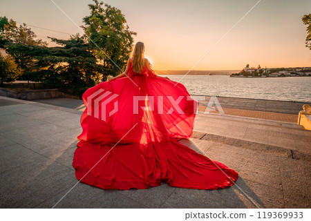 Sunrise red dress. A woman in a long red dress against the backdrop of sunrise, bright golden light of the sun's rays. The concept of femininity, harmony. 119369933
