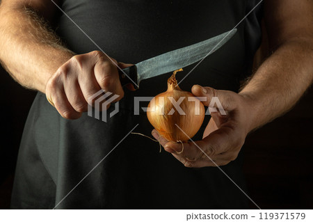 A cook tries to cut an onion with a knife. Low key concept of cooking a tasty and aromatic vegetable dish in the kitchen A cook tries to cut an onion with a knife. Low key concept of cooking a tasty and aromatic vegetable dish in the kitchen 119371579
