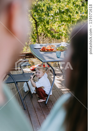 Curious toddler playing under table an outdoor family breakfast, with parents looking on lovingly Curious toddler playing under table an outdoor family breakfast, with parents looking on lovingly 119371618
