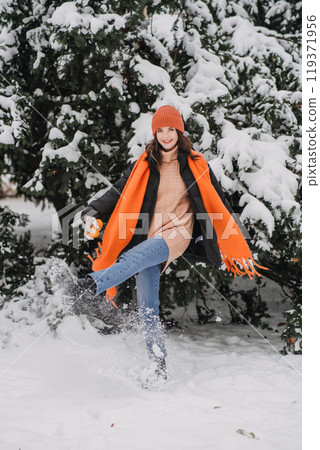 Woman in orange scarf and hat walking in snow, smiling and enjoying winter day outdoors. Winter fashion trends, nature exploration, Outdoor Winter Activities 119371956