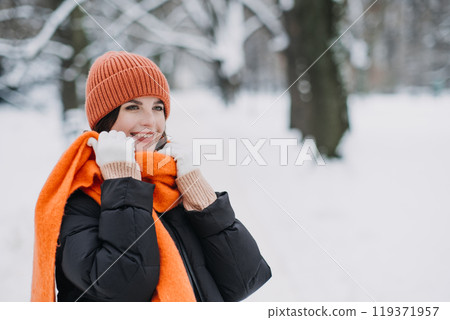 Woman in orange winter hat and scarf smiling in snow-covered park. Warm seasonal fashion, winter style trends, cozy outdoor clothing. Color Therapy in Seasonal Fashion 119371957