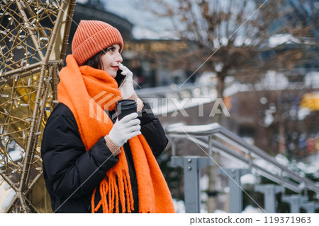 Millennial woman in cozy orange winter gear using smartphone. Christmas Urban tech-savvy style, winter warmth, city scene, hot coffee in hand. 119371963